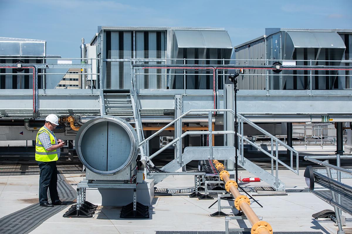 Mitie male employee, wearing a white hard hard and Mitie hi vis vest, inspecting large equipment on a roof